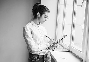 Young woman standing near window in office and writing ideas.