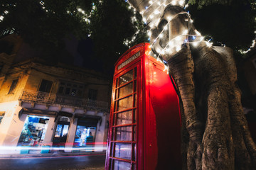 Red telephone box at night with light trails from cars and a tree with white fairy lights