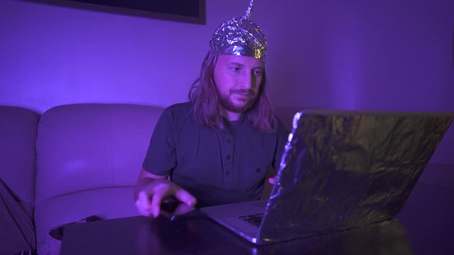 Man with long hair in Tin Foil Hat typing message on phone, then work at laptop covered with foil. Foil hat shields him from 5G waves, electromagnetic fields, mind control, mind reading, global conspi