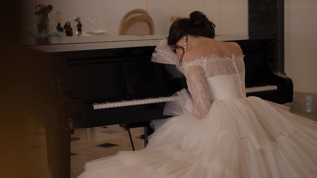 graceful girl in white dress with lacy bouffant skirt leans on piano in decorated wedding hall slow motion