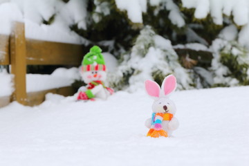 Felt figurines of a happy bunny and snowman on the background of a snowy pine tree and hedge.