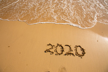 Summer beach holiday 2020 season golden sand - year - decade - new season lettering on the beach with wave and clear blue sea. Numbers 2020 year on the sea shore, message handwritten New Years concept