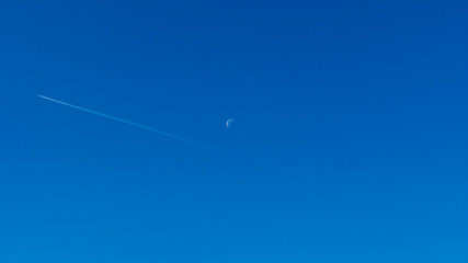 clear blue sky with the moon in the center of the screen and a plane flying near it. Wallpaper backgroun. 16x9 wide.