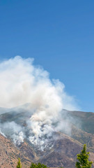 Vertical Mountain landscape with smoke from wild forest fire against clear blue sky