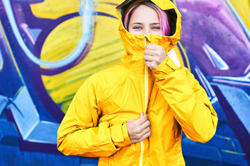 Urban bright portrait cool young woman in hood with cheerful smile wearing in yellow jacket posing against wall painted with graffiti. Urban clothing style, street style, lifestyle portrait.