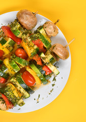 Vegetable skewers on a plate of mushrooms, tomatoes, zucchini, pepper, skewers, buttery, bright. Yellow background, vertical