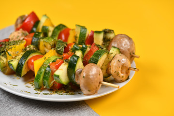 Vegetable skewers on a plate of mushrooms, tomatoes, zucchini, pepper, skewers, buttery, bright. Yellow background, horizontal
