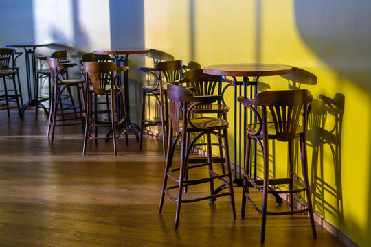 Tall antique wooden tables and chairs stand in a cafe, restaurant. A saturated shadow from sunlight is reflected in the foreground on the wall and floor, in the background is a cold shadow