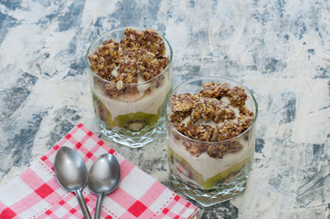 Dessert granola, Greek yogurt, kiwi and banana in two glass cups with spoons and pink checkered towel on gray concrete. Fitness, figure, body and healthy food
