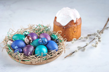 Dish with colored easter eggs.  The dish is stylized as a nest.  A pussy-willow branch and pastry made from yeast dough complete the composition.  Light background.  Close-up.  Free space for text.