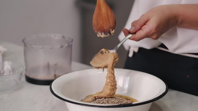 The chef is adding peanut butter to the mass. She is using a wooden spoon. The cook is preparing the filling for sweets.