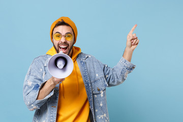Crazy young hipster guy in fashion jeans denim clothes posing isolated on pastel blue background in studio. People lifestyle concept. Mock up copy space. Scream in megaphone point index finger aside.