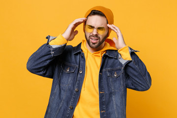 Tired young hipster guy in fashion jeans denim clothes posing isolated on yellow orange background. People lifestyle concept. Mock up copy space. Having headache put hands on head keeping eyes closed.