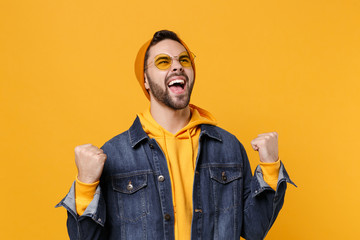 Joyful young hipster guy in fashion jeans denim clothes posing isolated on yellow orange background studio portrait. People emotions lifestyle concept. Mock up copy space. Doing winner gesture.