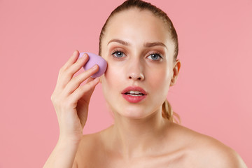 Obraz na płótnie Canvas Close up blonde half naked woman 20s perfect skin blue eyes isolated on pastel pink wall background studio portrait. Skin care healthcare procedures concept Apply foundation nude makeup sponge on face