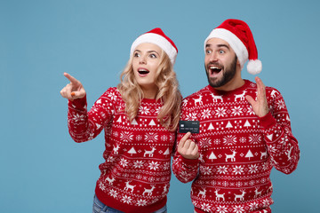 Amazed couple guy girl in Christmas sweater Santa hat posing isolated on blue background. New Year 2020 celebration party concept. Mock up copy space. Point index fingers aside hold credit bank card.