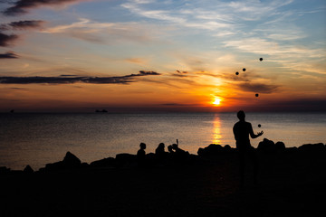 Juggler performance with many balls on the beach of the mediterranean sea in trieste, italy while sunset. Only silhouette visible at dusk. Couple and other people sitting at the coast line.
