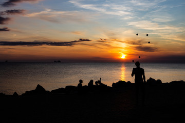 Juggler performance with many balls on the beach of the mediterranean sea in trieste, italy while sunset. Only silhouette visible at dusk. Couple and other people sitting at the coast line.
