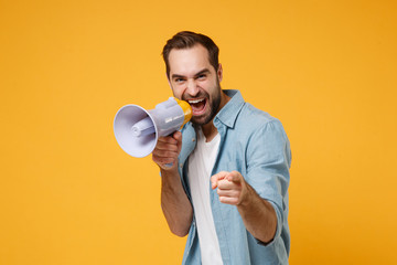 Funny young man in casual blue shirt posing isolated on yellow orange background studio portrait. People lifestyle concept. Mock up copy space. Screaming in megaphone pointing index finger on camera. - obrazy, fototapety, plakaty