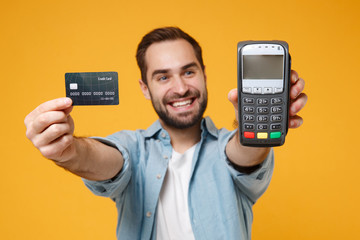Smiling young man in blue shirt posing isolated on yellow orange background. People lifestyle concept. Mock up copy space. Hold wireless bank payment terminal to process acquire credit card payments.