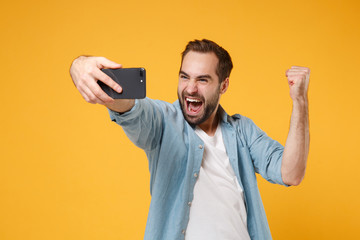 Happy young man in casual blue shirt posing isolated on yellow orange wall background. People lifestyle concept. Mock up copy space. Doing selfie shot on mobile phone, doing winner gesture, screaming.