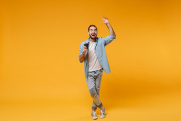 Fototapeta na wymiar Funny young man in casual blue shirt posing isolated on yellow orange background. People lifestyle concept. Mock up copy space. Hold cup of coffee or tea, waving greeting with hand as notices someone.