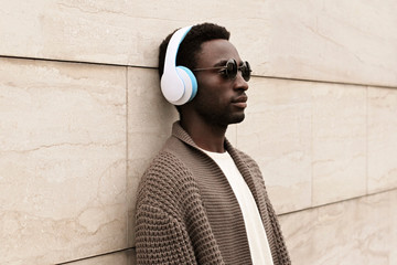 Portrait stylish african man in wireless headphones listening to music wearing brown knitted cardigan and sunglasses on city street over brick wall background
