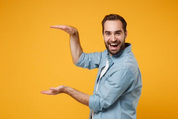 Cheerful young bearded man in casual blue shirt posing isolated on yellow orange wall background....