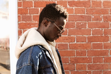 Portrait close up side view stylish african man wearing sunglasses and jeans jacket with hood on city street over brick textured wall background
