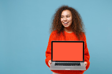 Funny young african american girl in casual orange knitted clothes isolated on pastel blue background. People lifestyle concept. Mock up copy space. Hold laptop pc computer with blank empty screen.