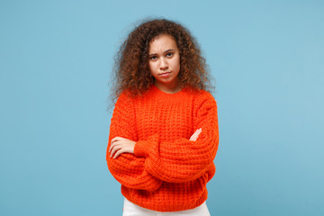 Dissatisfied young african american girl in casual orange knitted clothes isolated on pastel blue wall background studio portrait. People lifestyle concept. Mock up copy space. Holding hands crossed.
