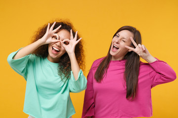 Two women friends european african girls in pink green clothes isolated on yellow background. People lifestyle concept. Mock up copy space. Showing victory sign imitating glasses with hands near eyes.