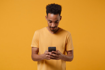 Young african american guy in casual t-shirt posing isolated on yellow orange wall background studio portrait. People lifestyle concept. Mock up copy space. Using mobile phone, typing sms message.