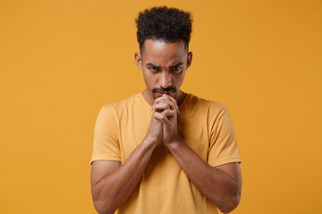 Young african american guy in casual t-shirt posing isolated on yellow orange wall background studio portrait. People emotions lifestyle concept. Mock up copy space. Holding hands folded in prayer.