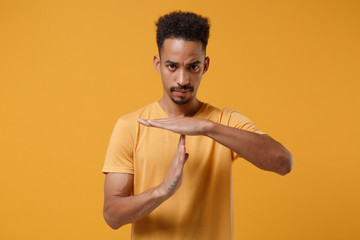 Serious young african american guy in casual t-shirt posing isolated on yellow orange background studio portrait. People emotions lifestyle concept. Mock up copy space. Hold hands perpendicularly. - obrazy, fototapety, plakaty