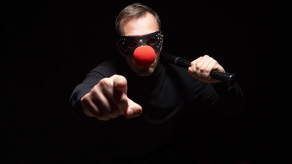 man in a leather mask with a red clown nose waving a bat on a black background