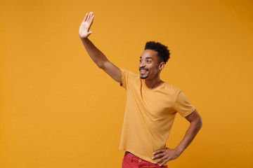 Pleasant young african american guy in casual t-shirt posing isolated on yellow orange wall background. People lifestyle concept. Mock up copy space. Waving and greeting with hand as notices someone.