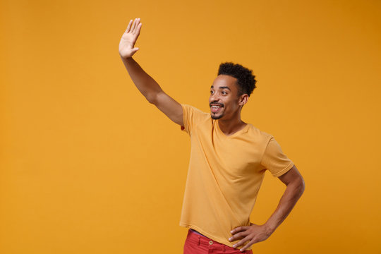 Pleasant young african american guy in casual t-shirt posing isolated on yellow orange wall background. People lifestyle concept. Mock up copy space. Waving and greeting with hand as notices someone.
