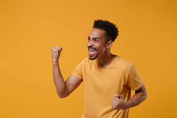 Happy young african american guy in casual t-shirt posing isolated on yellow orange wall background studio portrait. People emotions lifestyle concept. Mock up copy space. Clenching fists like winner.