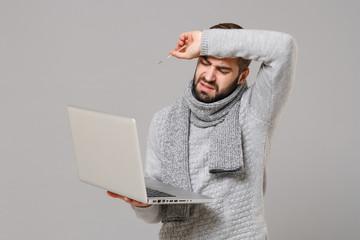 Man in gray sweater, scarf isolated on grey background. Healthy lifestyle, ill sick disease, online treatment consulting, cold season concept. Mock up copy space. Hold laptop pc computer, thermometer.