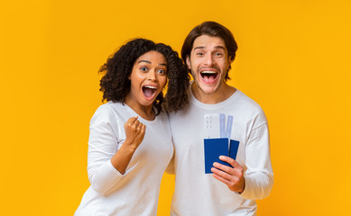 Happy interracial couple holding passports with tickets, posing over yellow background