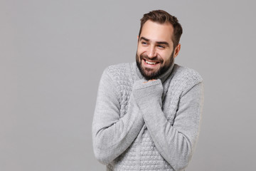 Smiling young man in gray sweater posing isolated on grey background, studio portrait. Healthy fashion lifestyle, people emotions cold season concept. Mock up copy space. Warming hands, looking aside.