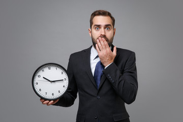 Shocked young bearded business man in classic suit shirt tie posing isolated on grey background. Achievement career wealth business concept. Mock up copy space. Holding clock covering mouth with hand.