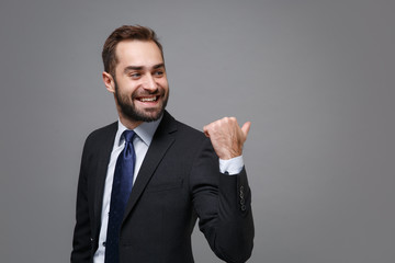 Smiling young bearded business man in classic black suit shirt tie posing isolated on grey background in studio. Achievement career wealth business concept. Mock up copy space. Pointing thumb aside.