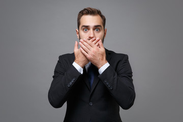 Shocked young bearded business man in classic black suit shirt tie posing isolated on grey wall background. Achievement career wealth business concept. Mock up copy space. Covering mouth with hands.