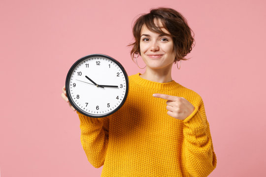 Smiling young brunette woman girl in yellow sweater posing isolated on pastel pink background in studio. People sincere emotions lifestyle concept. Mock up copy space. Pointing index finger on clock.