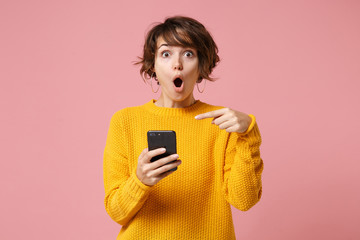 Shocked young brunette woman girl in yellow sweater posing isolated on pastel pink wall background studio portait. People lifestyle concept. Mock up copy space. Pointing index finger on mobile phone.