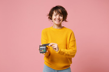 Young brunette woman in yellow sweater posing isolated on pink background. People lifestyle concept. Mock up copy space. Hold bank payment terminal to process, acquire credit card payments, blinking. - obrazy, fototapety, plakaty