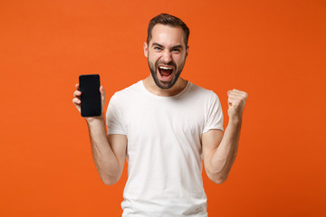 Crazy young man in casual white t-shirt posing isolated on orange wall background. People lifestyle concept. Mock up copy space. Holding mobile phone with blank empty screen, doing winner gesture.