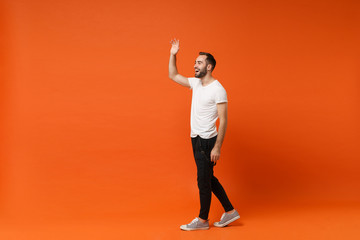 Fototapeta na wymiar Side view of cheerful young man in casual white t-shirt posing isolated on orange background in studio. People lifestyle concept. Mock up copy space. Waving and greeting with hand as notices someone.